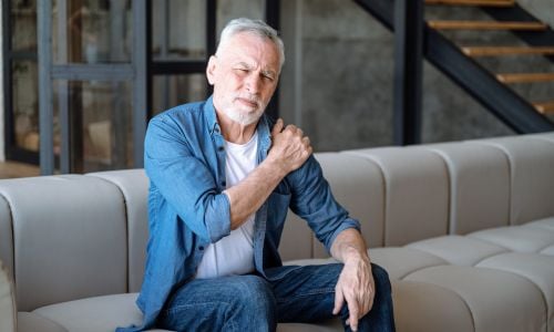 Uomo adulto seduto con il divano che si tocca la spalla a causa di dolori alle ossa e stanchezza