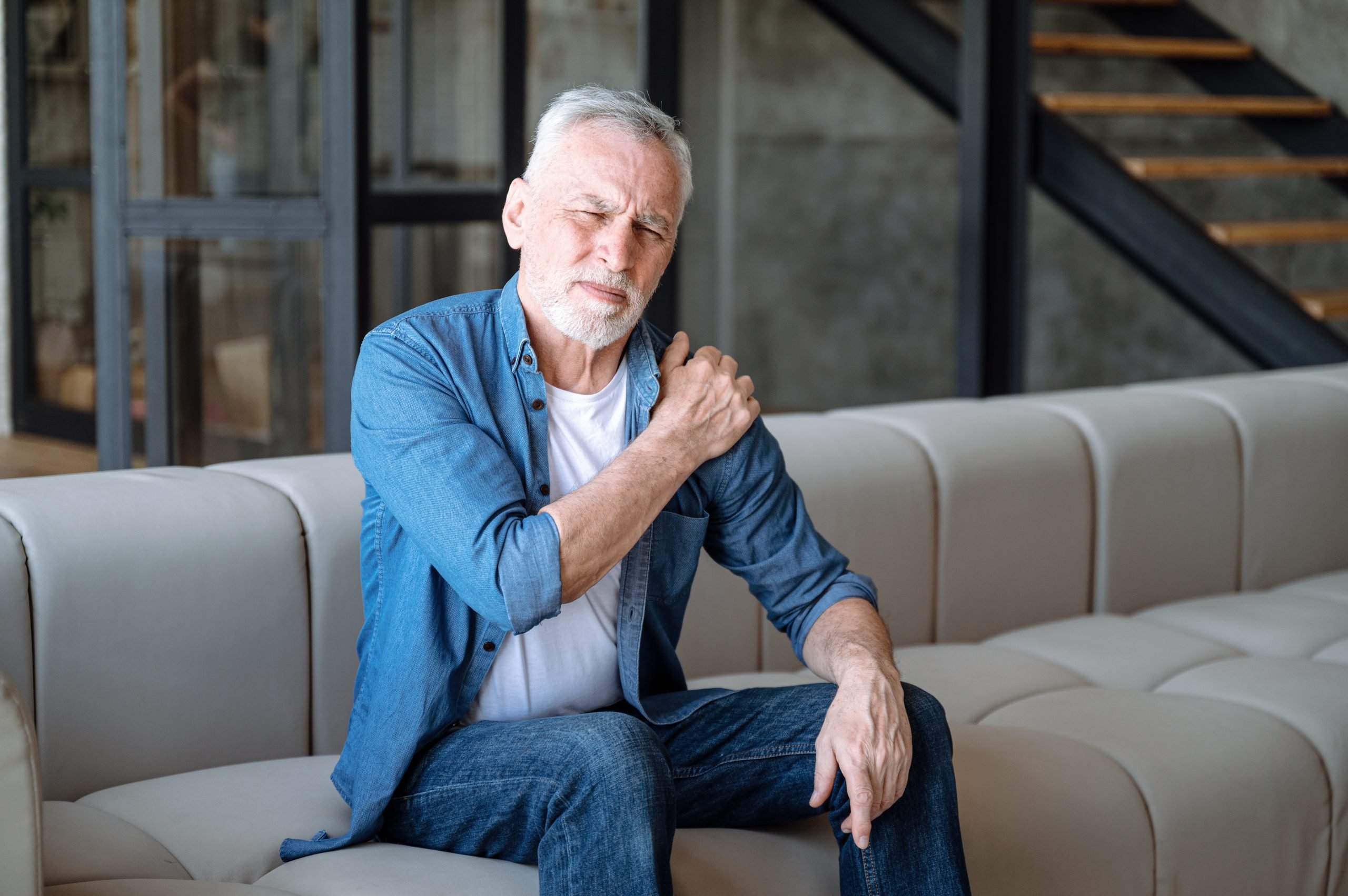 Uomo adulto seduto con il divano che si tocca la spalla a causa di dolori alle ossa e stanchezza